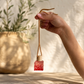 Hand holding a small red and gold decorative air freshener bottle with a blurred natural background